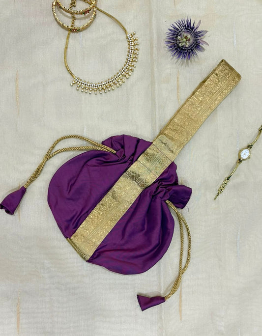 A purple drawstring pouch with a wide gold zari border, laid flat on a light beige fabric background alongside jewelry accessories.