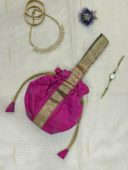 A magenta pink fabric pouch bag with a gold zari embroidered strap and drawstring tassels, styled on a cream-colored fabric background alongside gold jewelry and a watch.