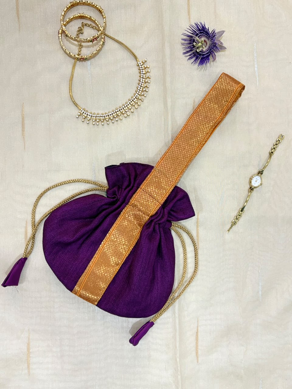 A purple drawstring pouch with a wide gold zari strap, styled on a cream fabric background with gold jewelry and a small purple flower.