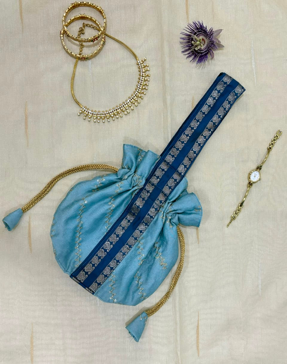 A light blue traditional drawstring pouch with a dark blue embroidered border and gold tassel ties, displayed on a cream-colored textured fabric alongside gold jewelry accessories.
