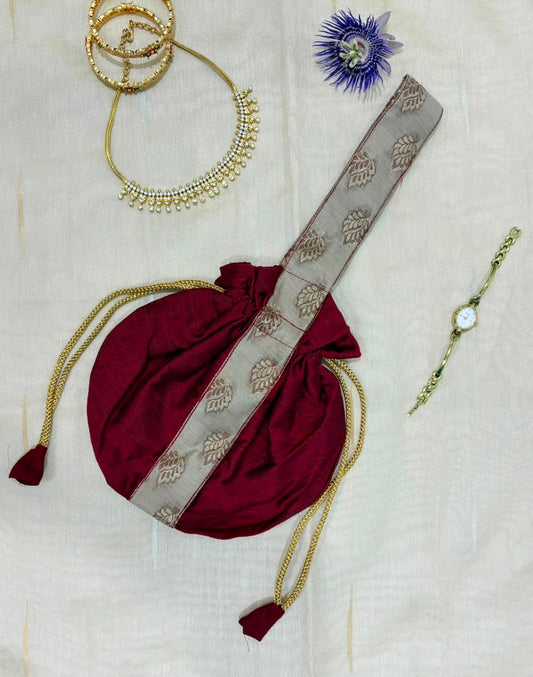 A maroon drawstring pouch with a beige patterned strap, styled on a white fabric background with gold jewelry accessories including necklaces, a bracelet, and a blue flower.