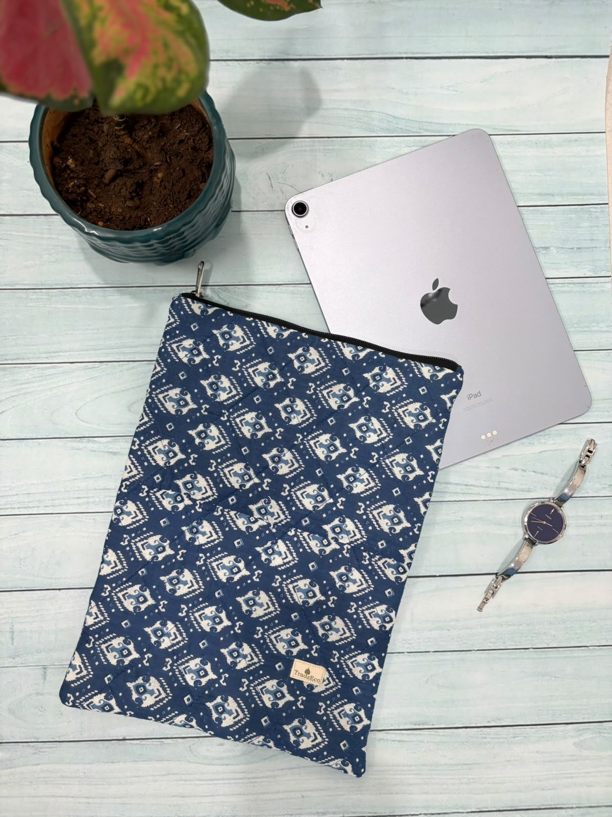 A navy blue and white patterned tablet sleeve is laid flat on a light wood surface, styled with a silver tablet, a potted plant, and a watch.
