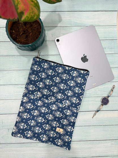 A navy blue and white patterned tablet sleeve is laid flat on a light wood surface, styled with a silver tablet, a potted plant, and a watch.