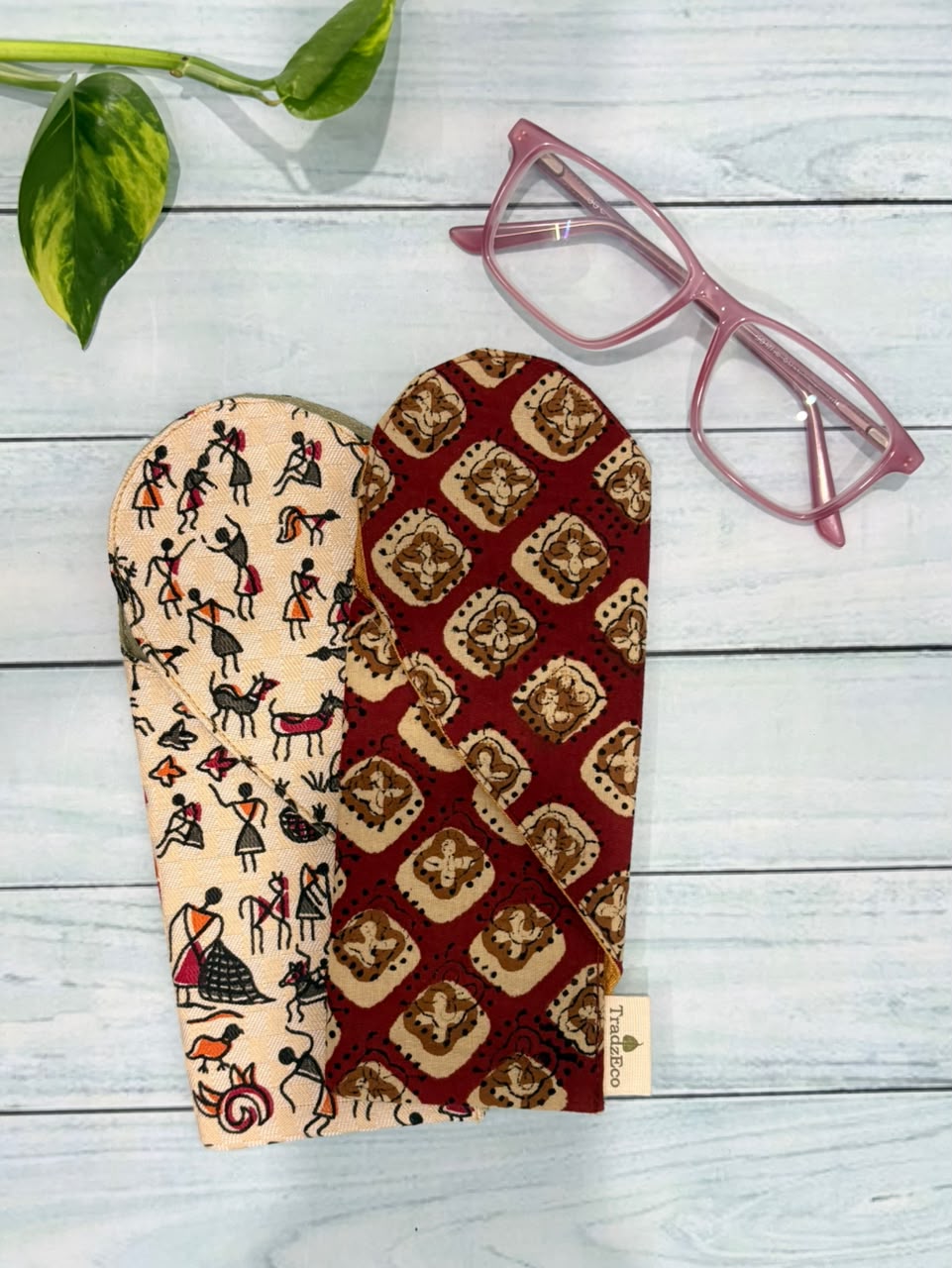 Two fabric eyeglasses pouches displayed on a light gray wood plank background, alongside pink eyeglasses and a green plant stem. One pouch is cream with black folk-art style figures, and the other is dark red with a gold geometric pattern.